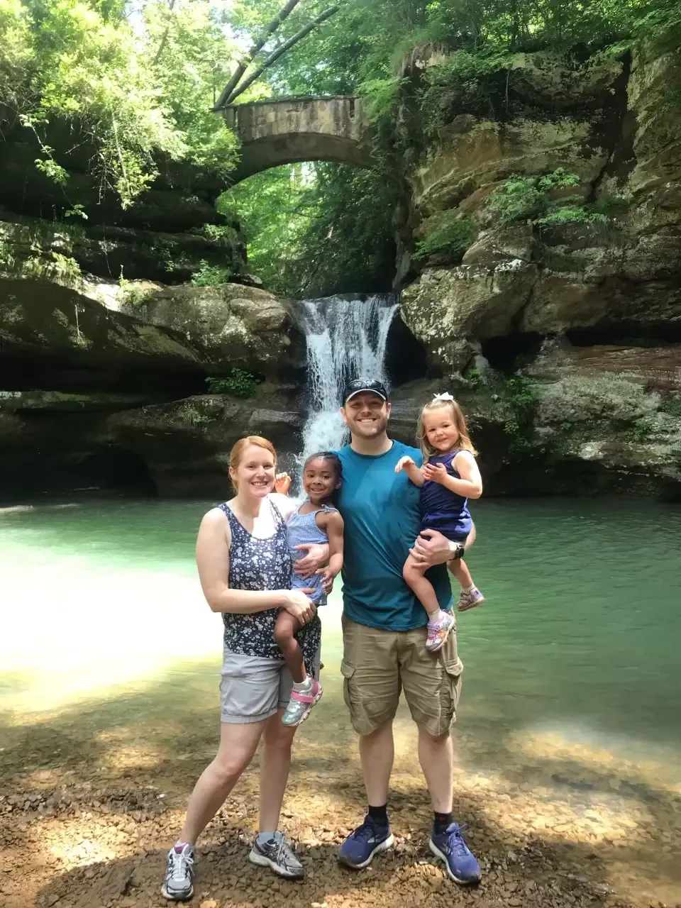 Family of four stands in front of a waterfall, bridge overhead, with a green/blue pool of water.