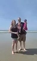 Family of four on a beach; mother, father holding a baby, and a child stands next to them on a sunny day.