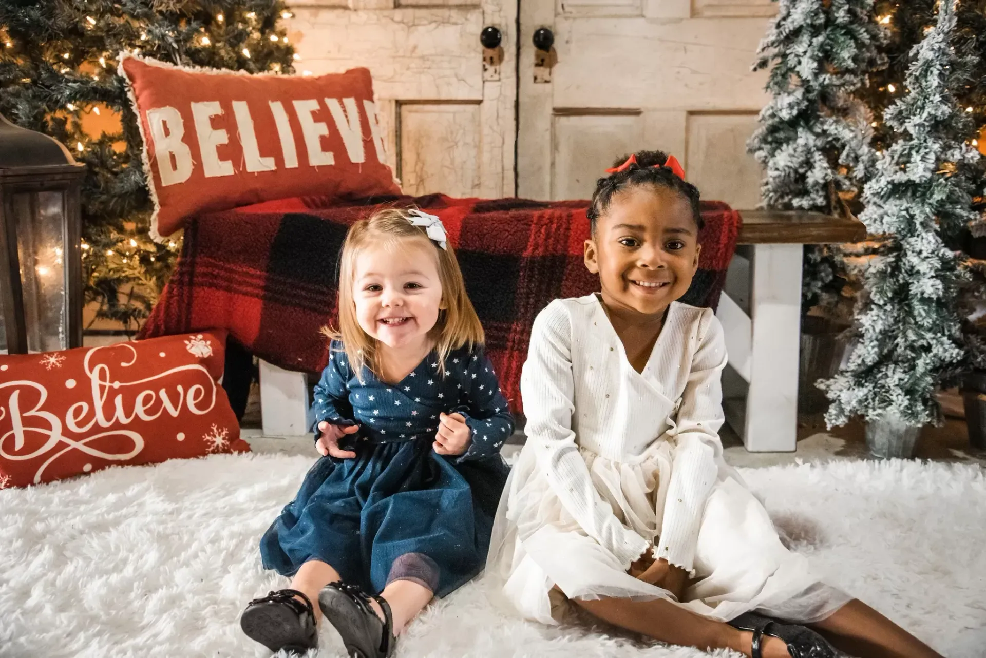 Two young girls smiling and seated on a fluffy white rug, with holiday decorations in the background.