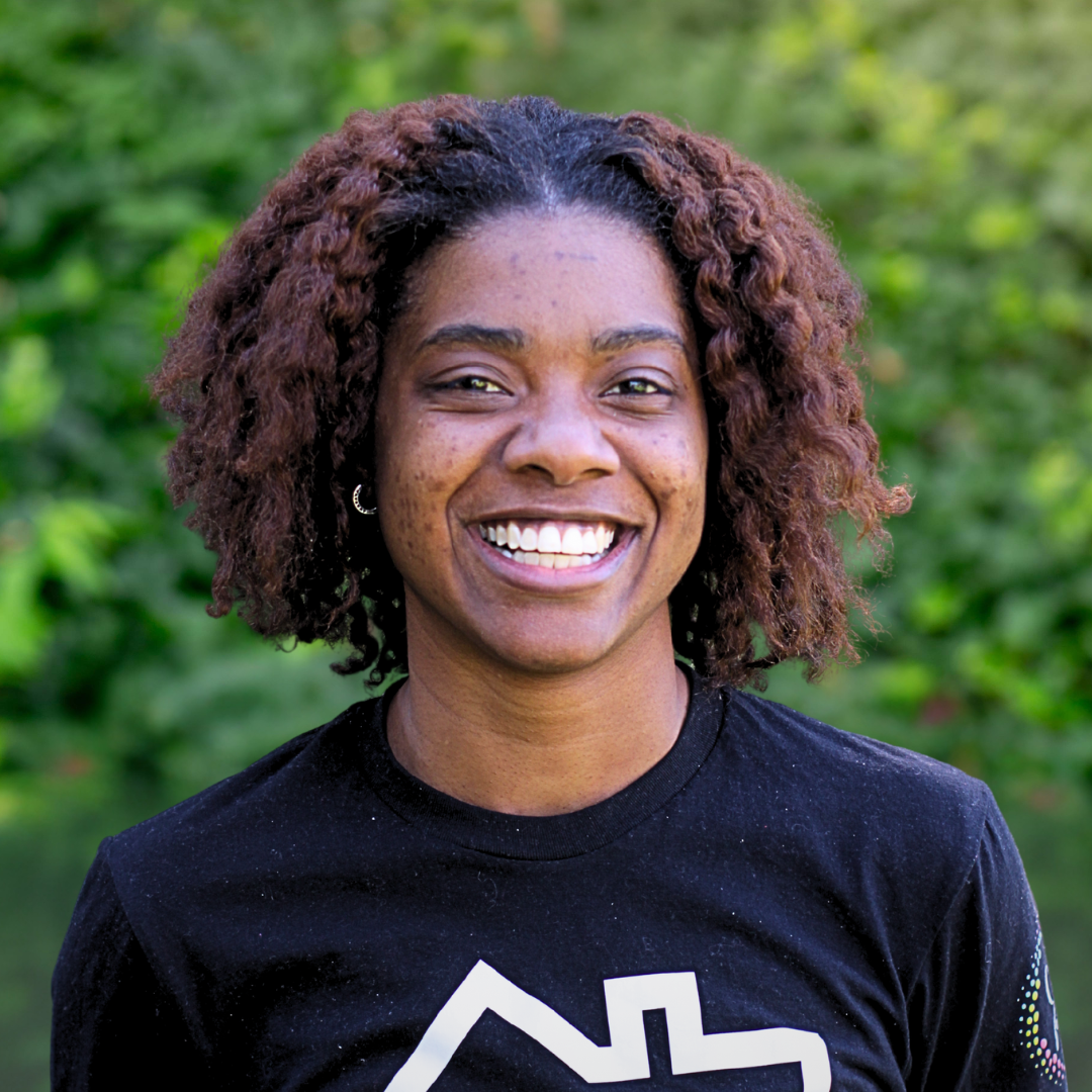 Smiling person with curly hair wearing a black shirt with a white logo, standing outdoors in front of greenery