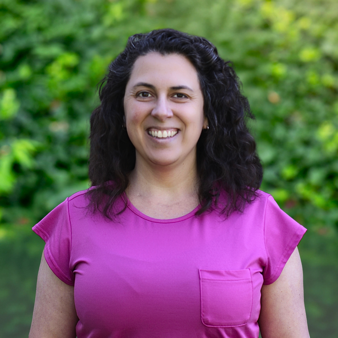 Woman with curly brown hair smiling, wearing a pink shirt, against a gray background.