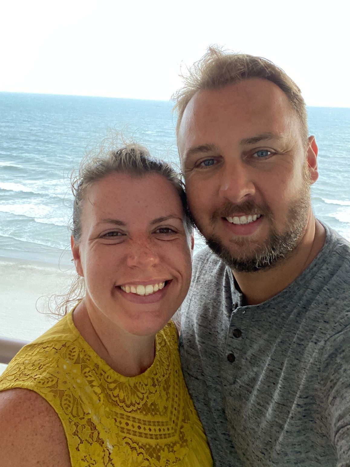 Couple smiling in front of the ocean. Woman in yellow top, man in gray shirt.