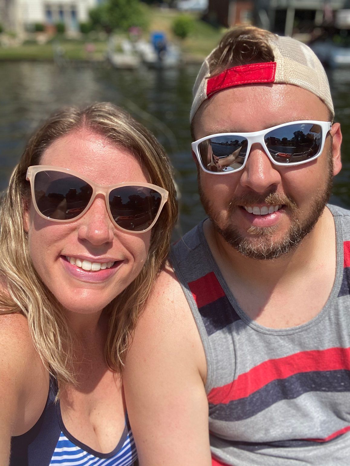 Couple smiles at the camera, boating. Woman wears gold glasses, man in a red-striped tank top and hat, with white sunglasses.