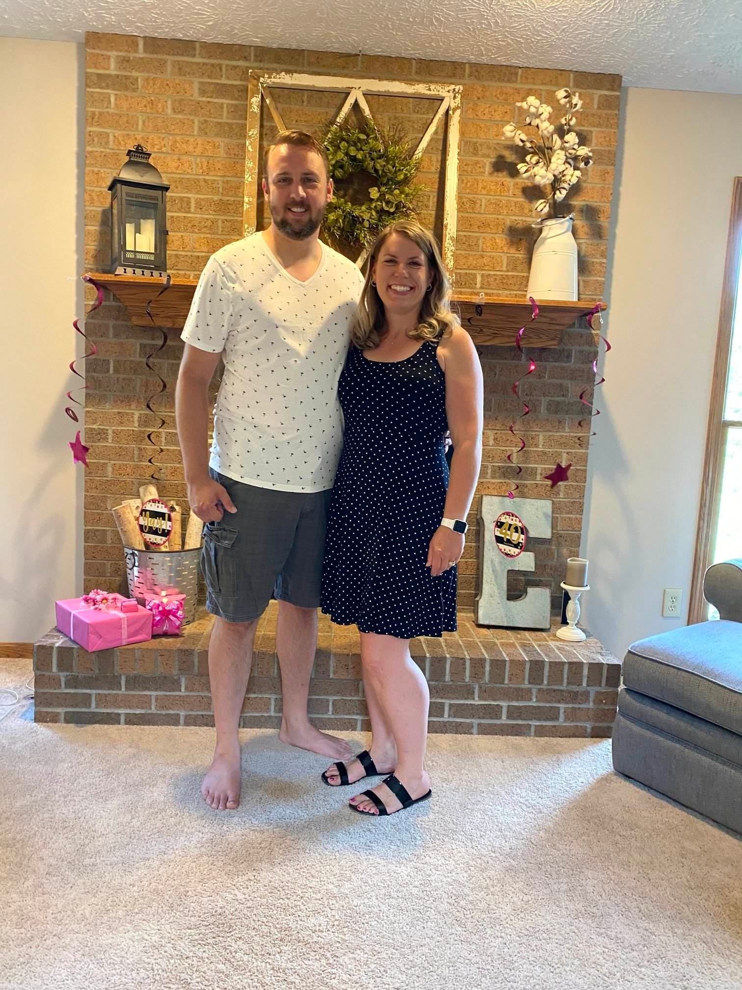 Couple poses in front of a brick fireplace; woman in polka-dot dress and sandals, man in shorts and dotted shirt.