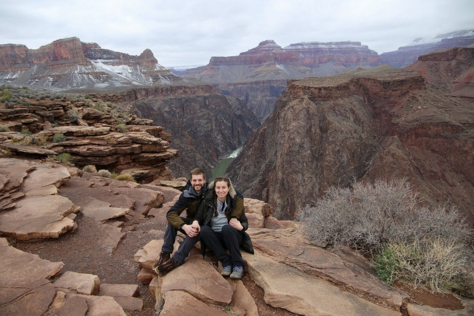 Couple seated on a rocky ledge overlooking the Grand Canyon. Snowy peaks, brown canyon walls, cloudy sky.