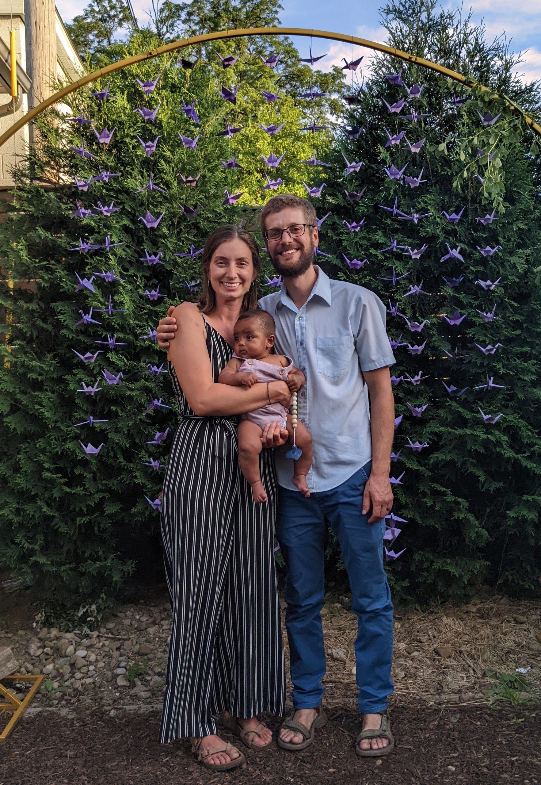Family of three poses outdoors: woman in black and white dress, man in blue shirt, child in arms.