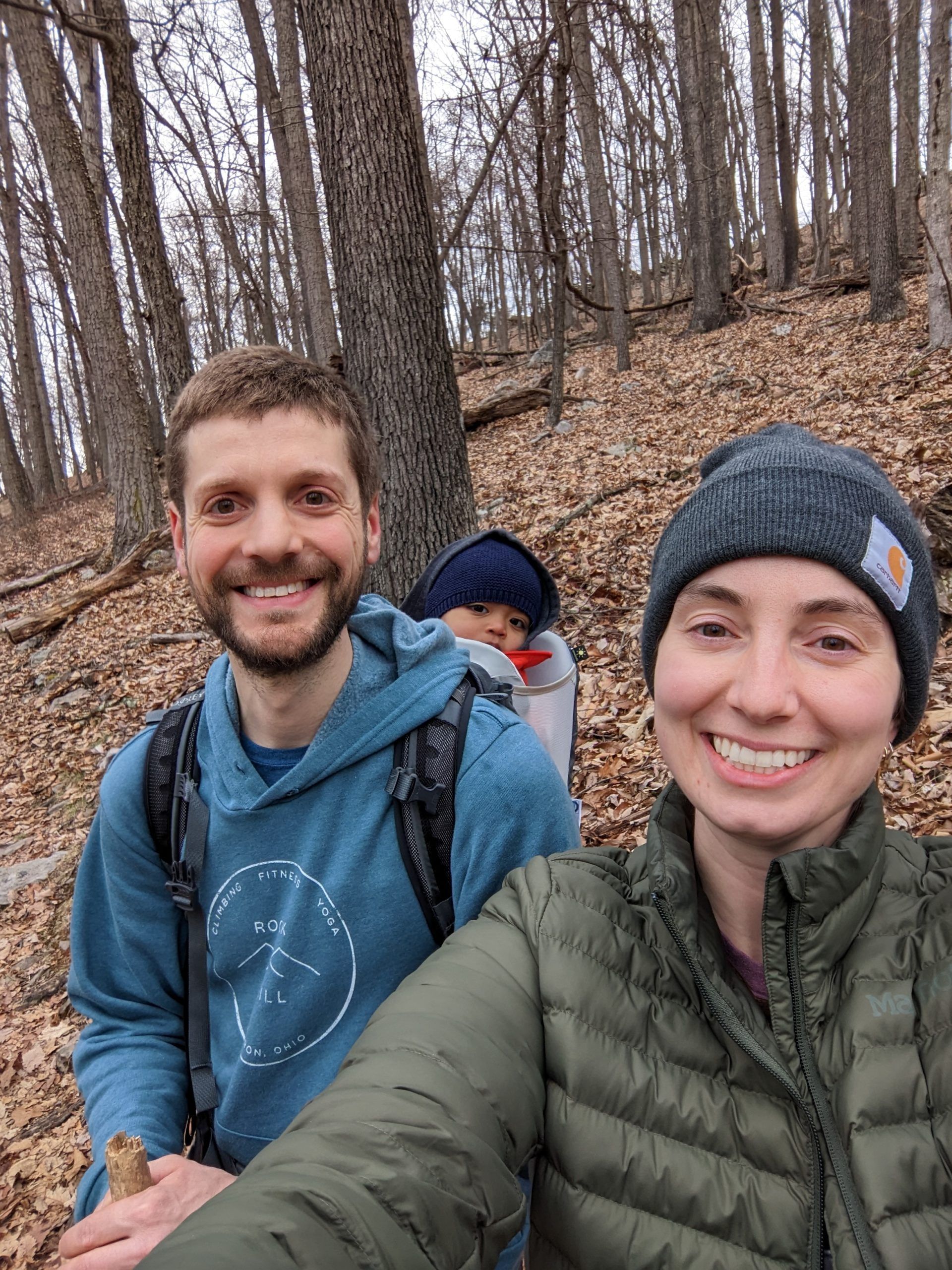Family hiking: man carries child, woman smiles, surrounded by trees.