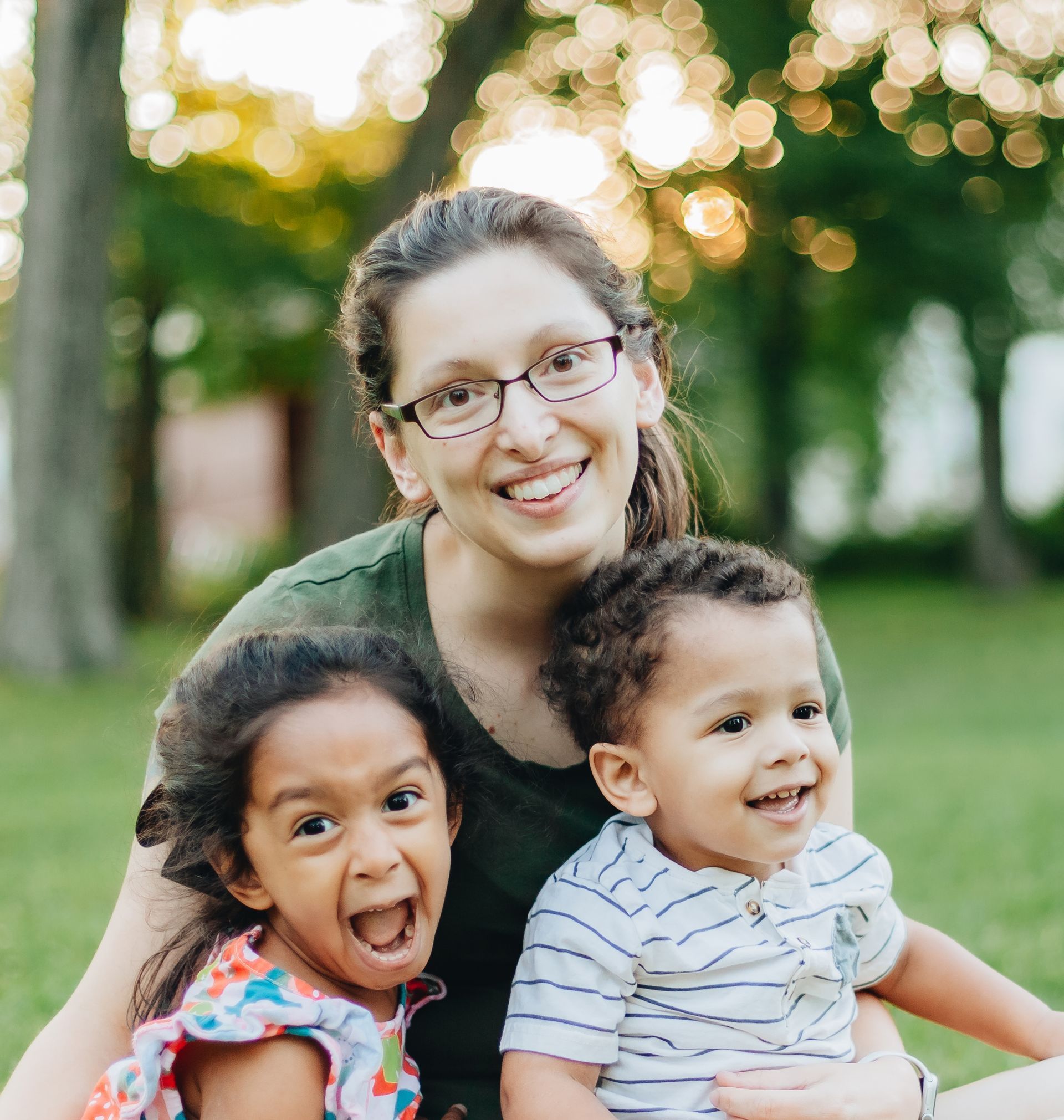 Woman with glasses smiles, holding two laughing children outdoors.