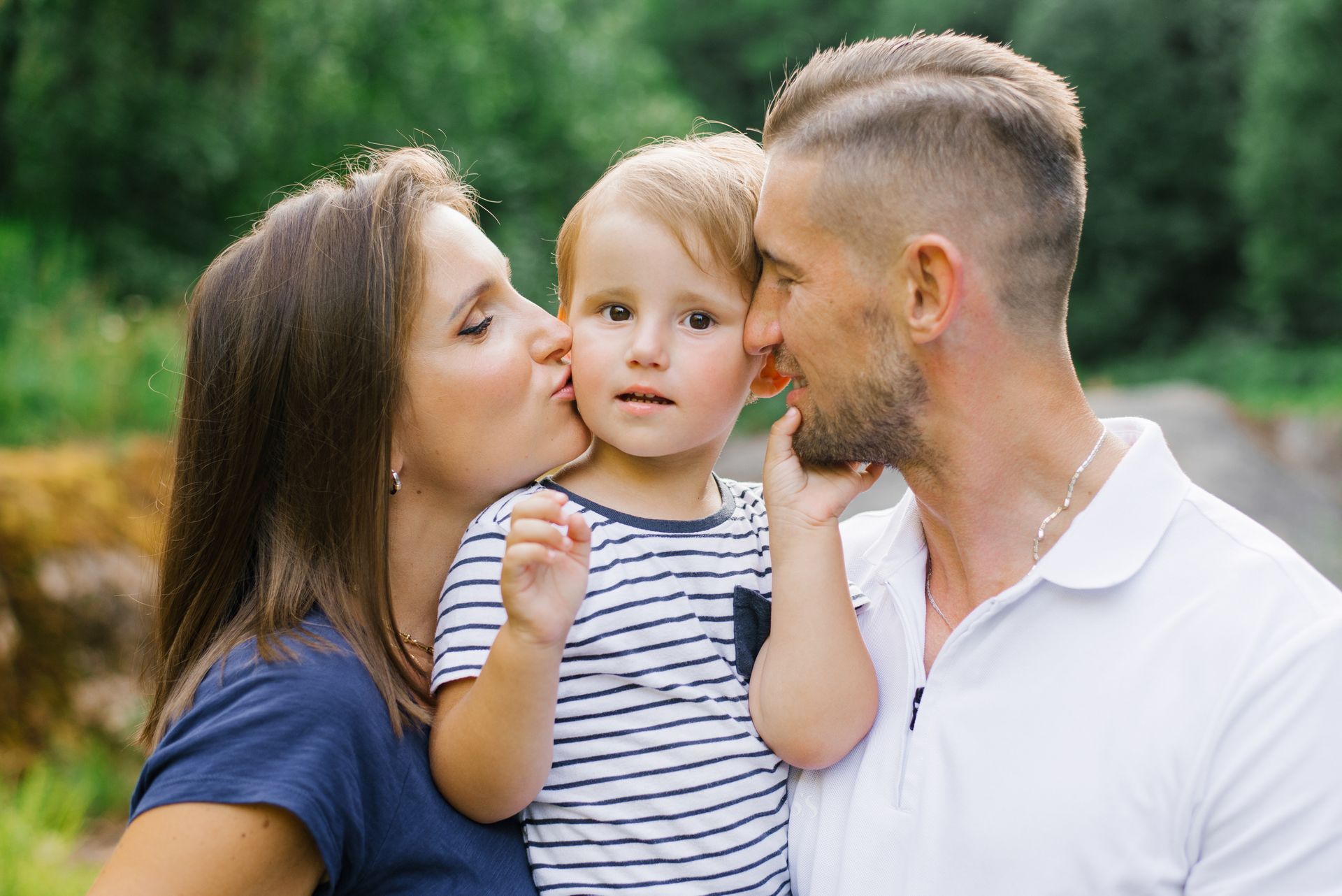 Family of three: woman kissing child's cheek, man holding child, outdoors with green foliage.