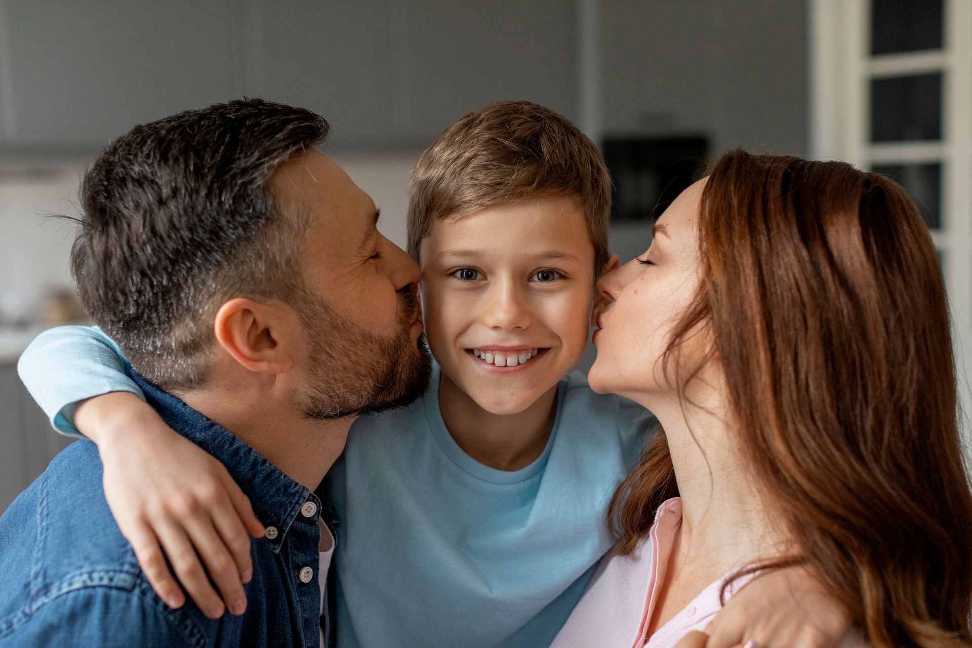 Parents kissing a smiling child on each cheek indoors.
