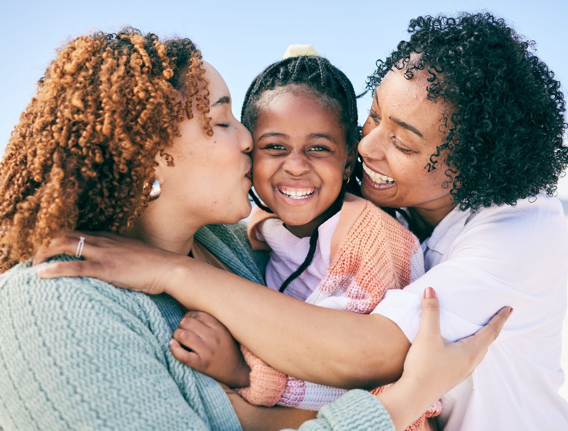Two women hugging a smiling child. One woman kisses child's cheek. All are outdoors, smiling.