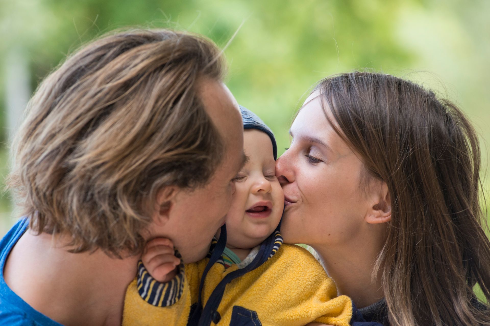 Parents kissing baby's cheeks outdoors; baby's face squished, wearing a blue hat and yellow jacket.