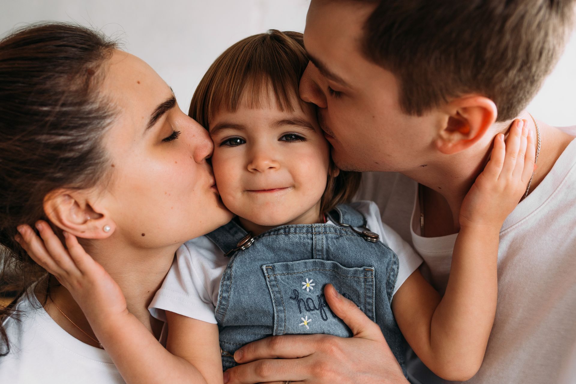 Parents kissing a child's cheeks, all smiling, in a bright setting. The child wears denim overalls.