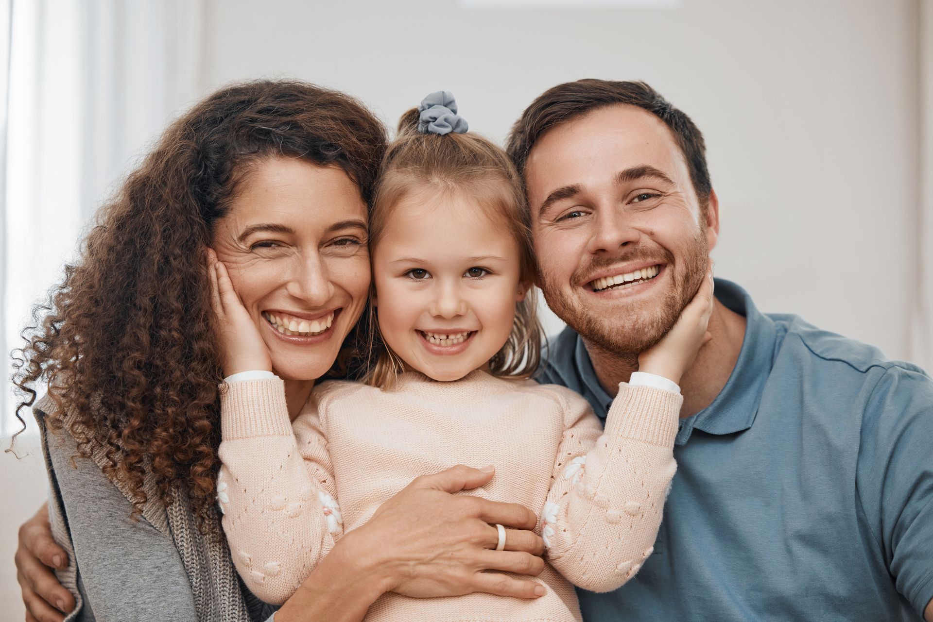 Family of three smiling, embracing. Woman on the left with curly hair, man on the right, child in the middle.