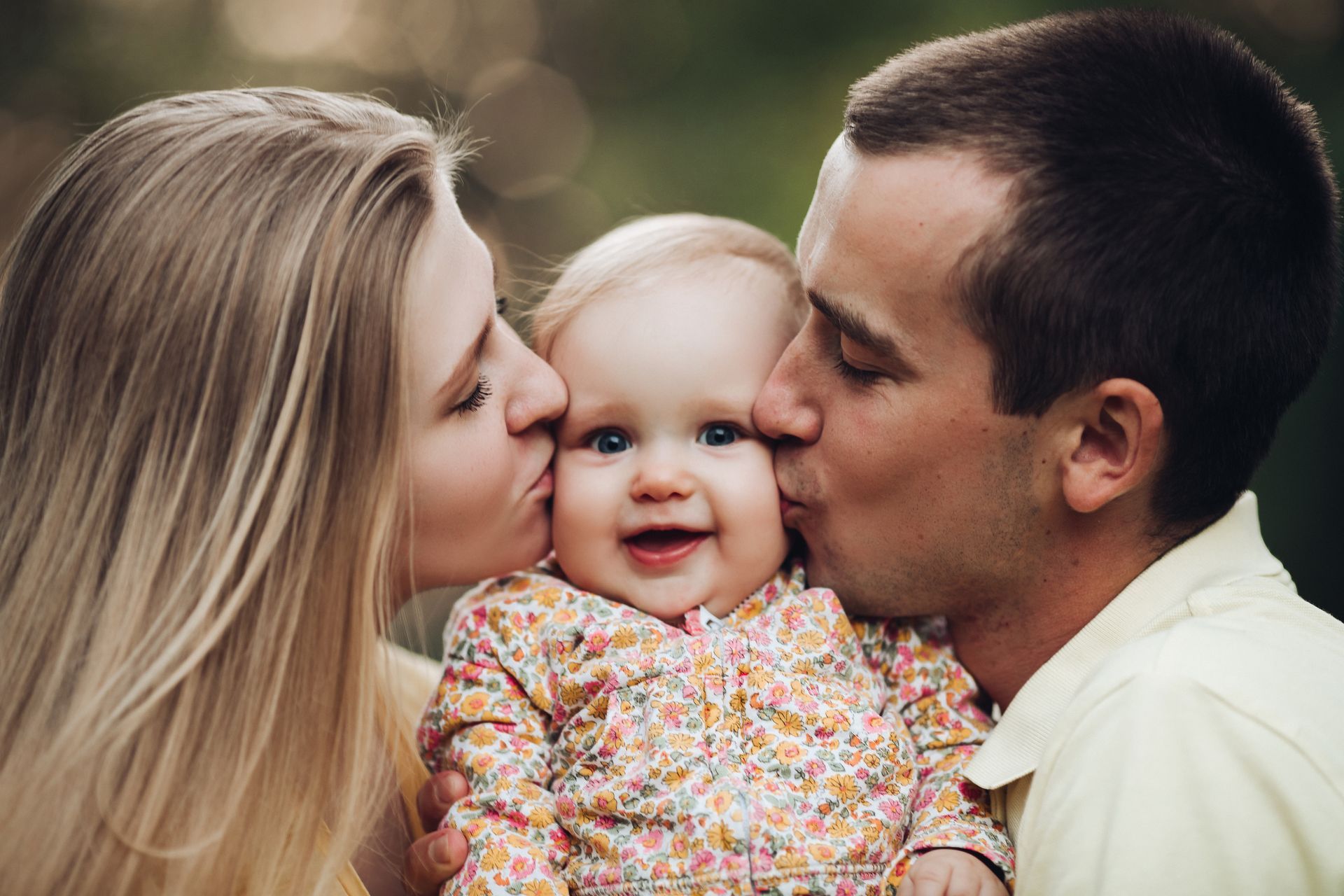 Parents kissing a smiling baby; outdoors, natural light.