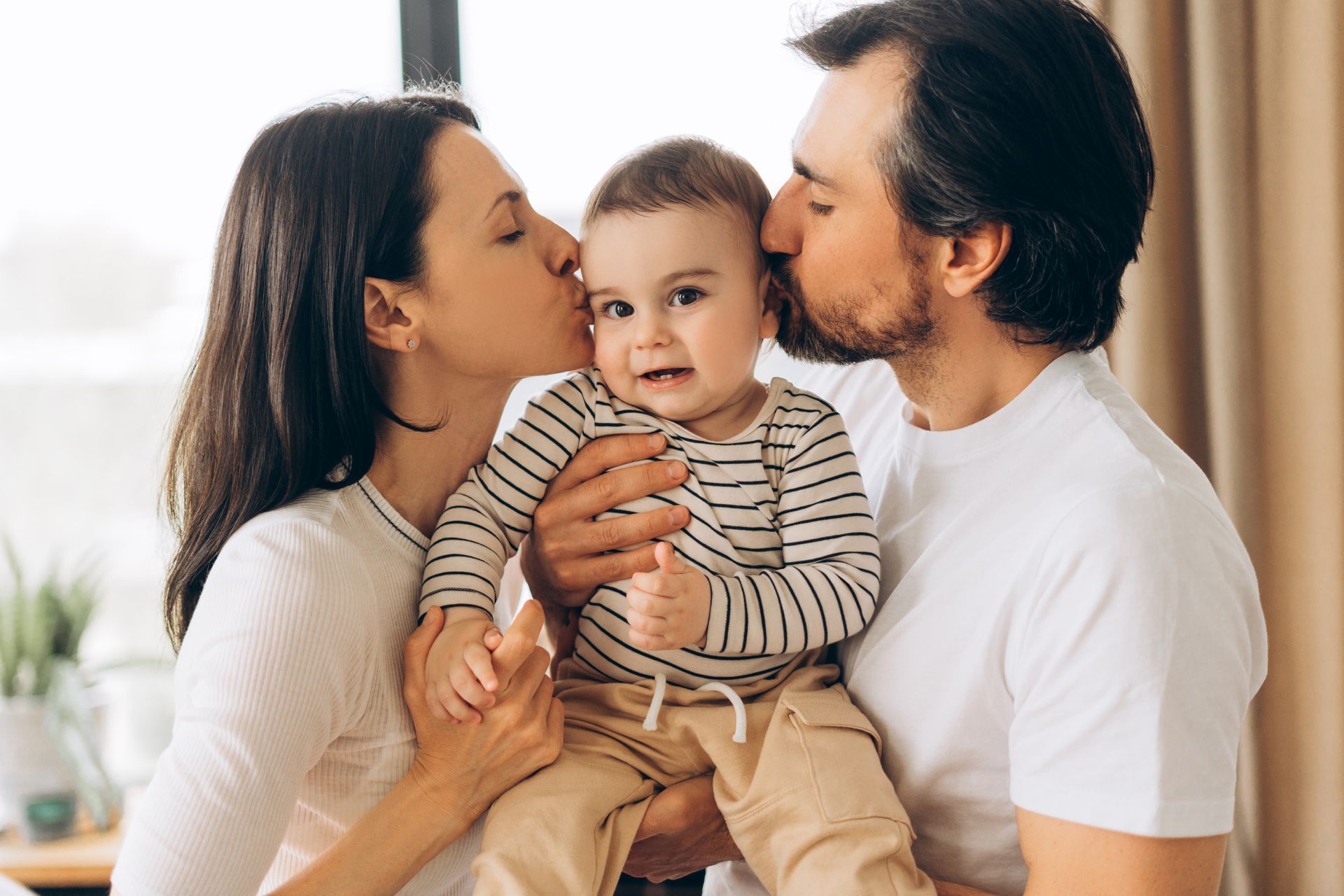 Parents kissing a baby on the cheeks. The baby is held between them, smiling.