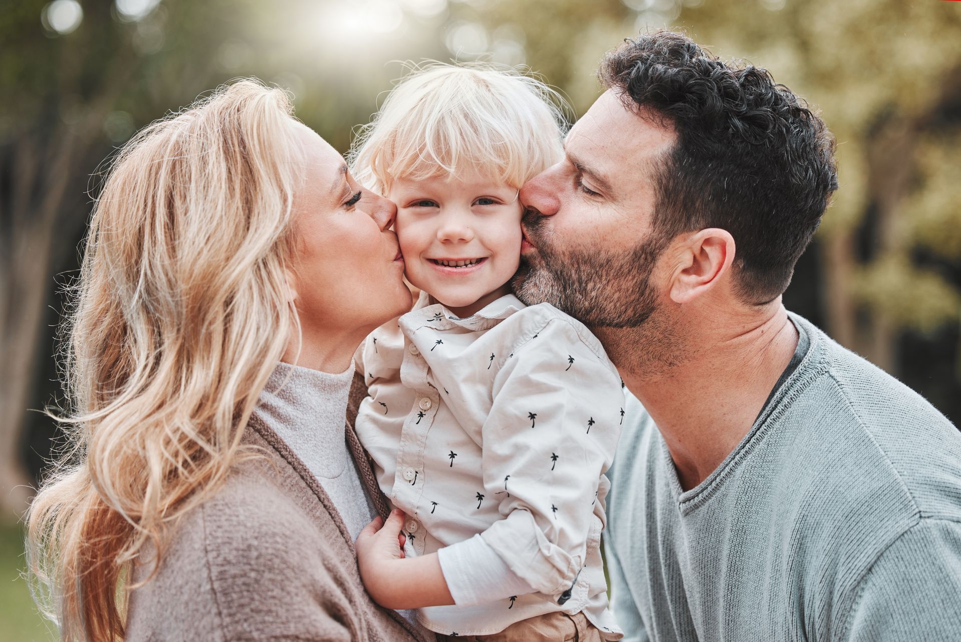 A child smiles as parents kiss his cheeks outdoors; light-haired individuals, trees in background.