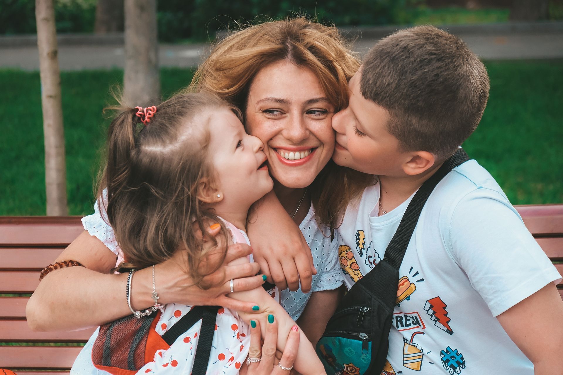 Woman smiles, embraced by children kissing her cheeks on a park bench.