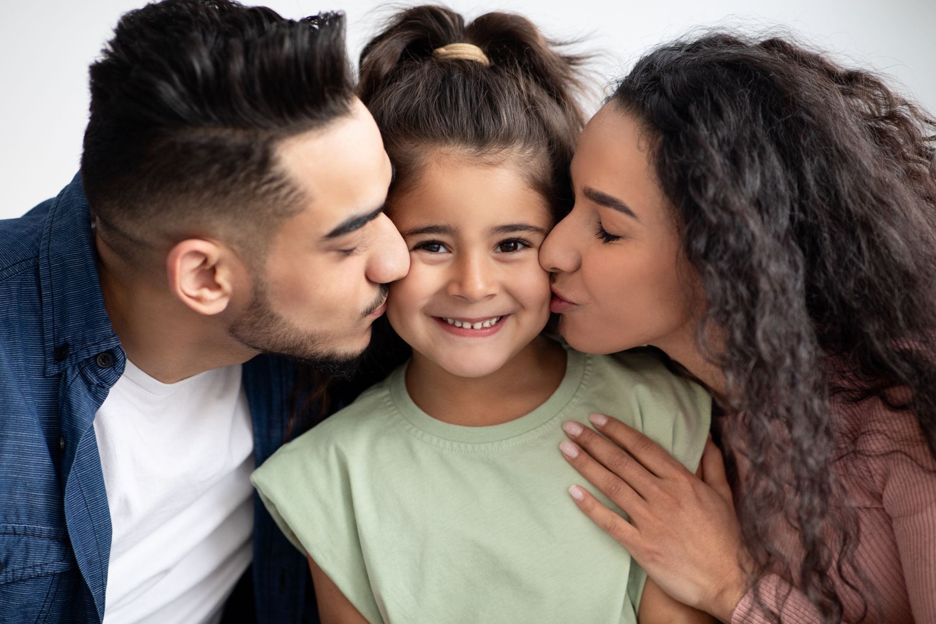 Parents kissing a smiling child on the cheeks, against a white background.