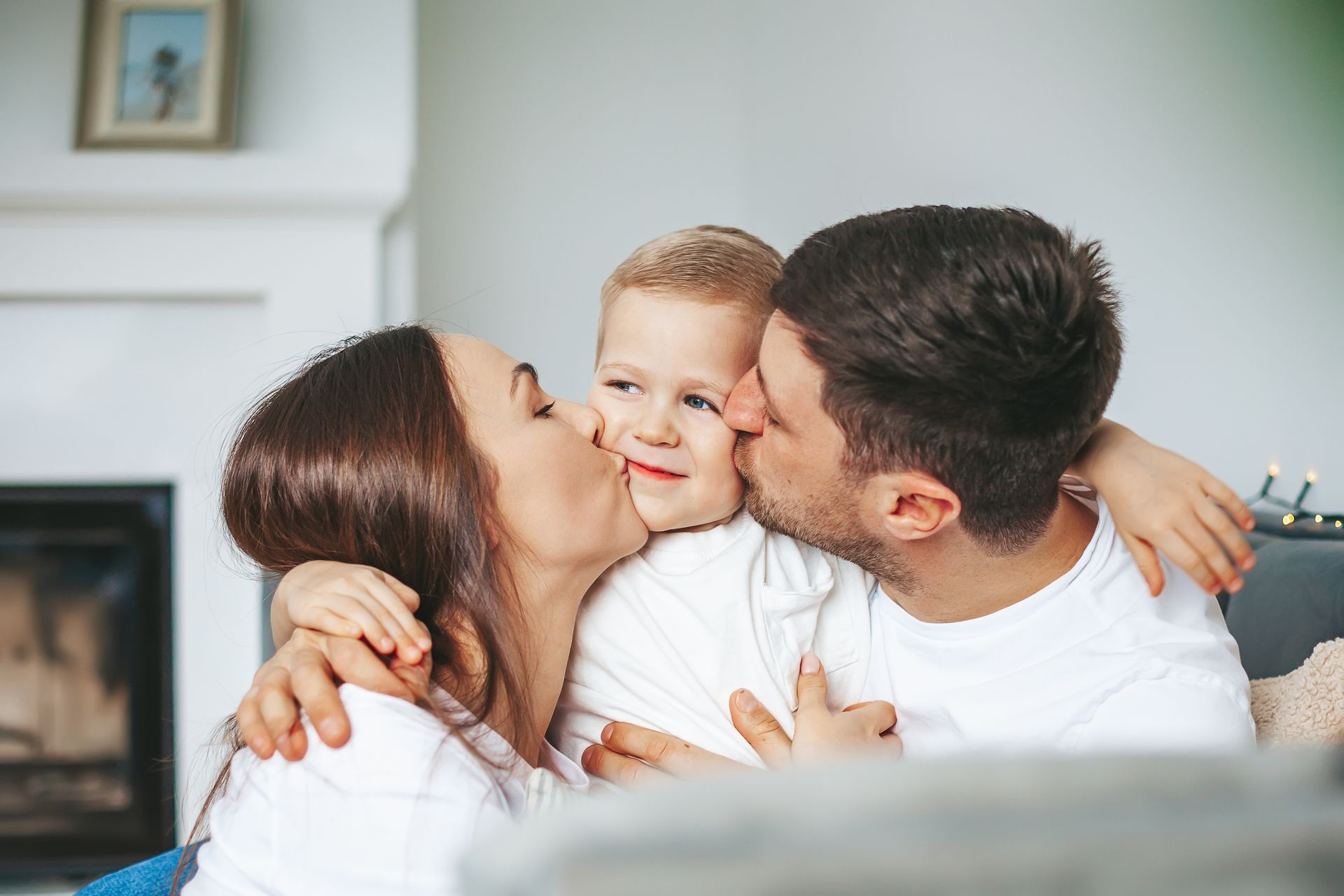 Parents kissing a child's cheeks, smiling. They are indoors near a fireplace.