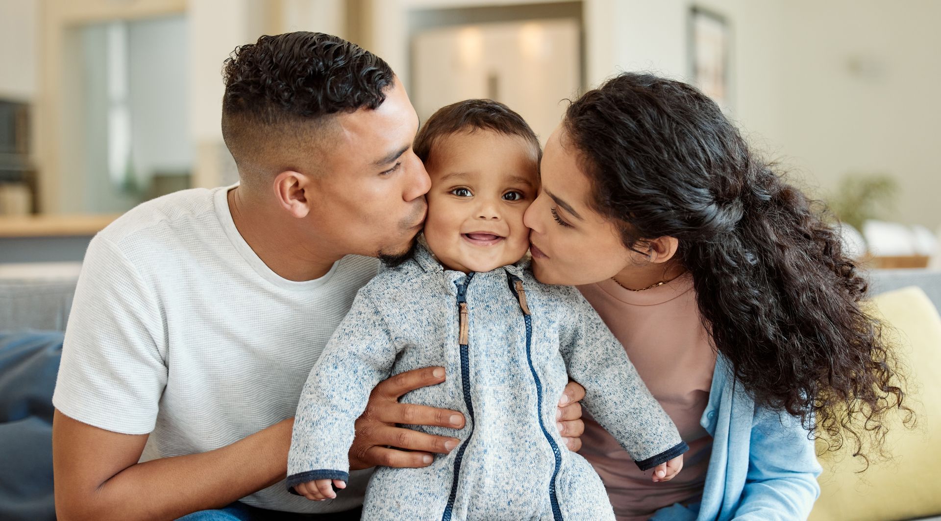 Parents kissing a smiling baby on the cheeks, sitting on a couch.