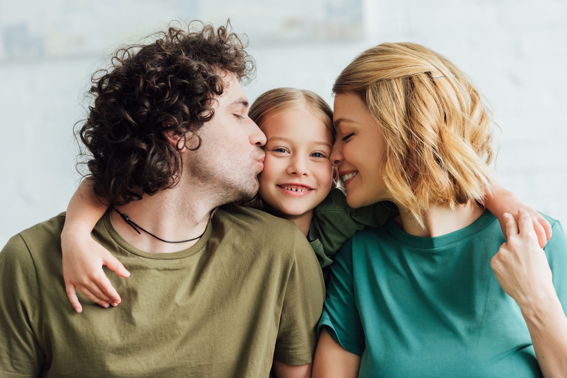 Family, with parents kissing a child's cheeks; all smiling, in a bright setting.