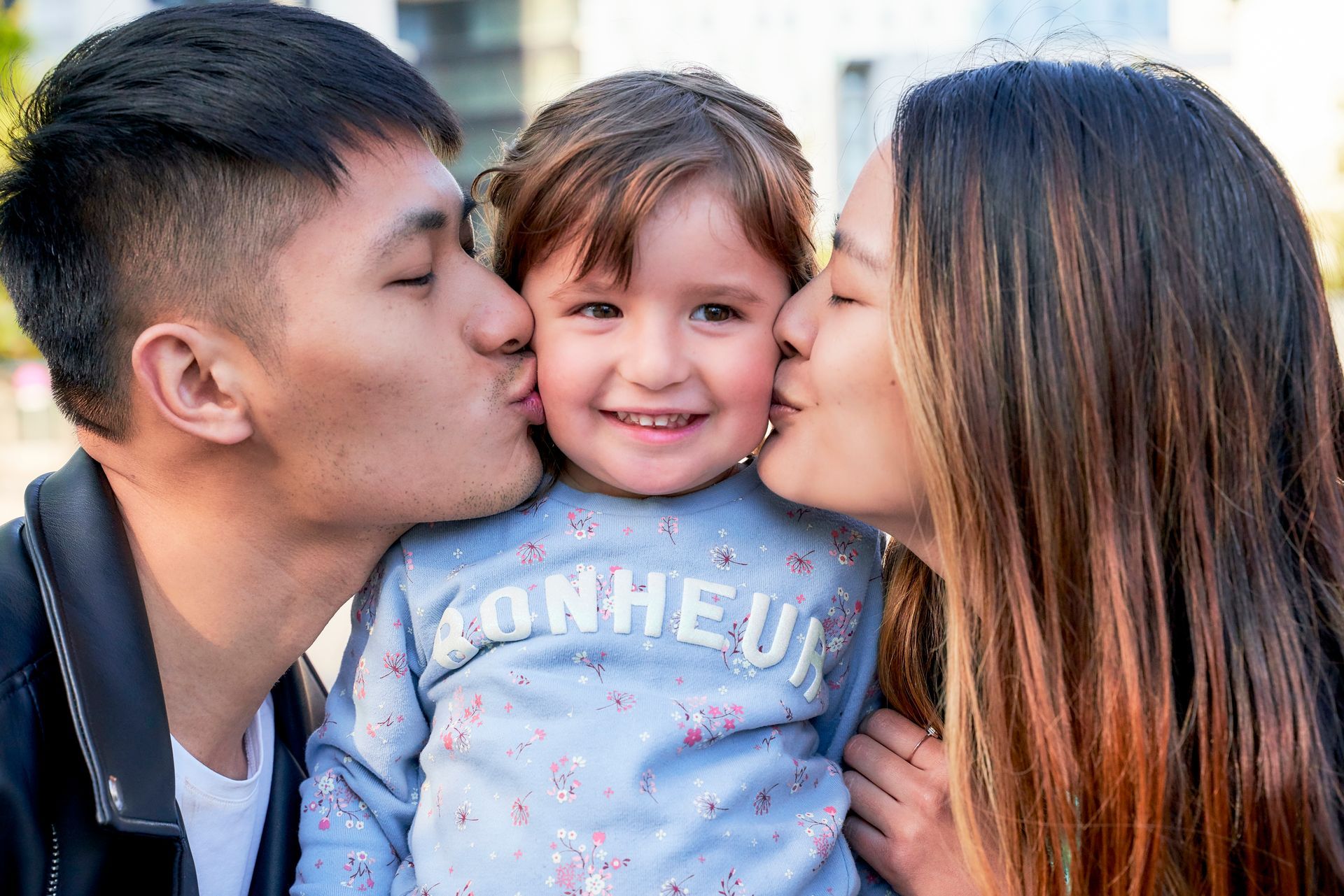 Parents kissing a smiling child on each cheek outdoors.