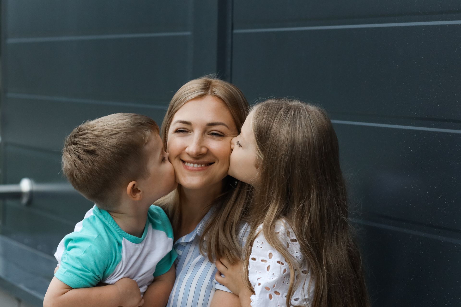 Woman smiles as two children kiss her cheeks.