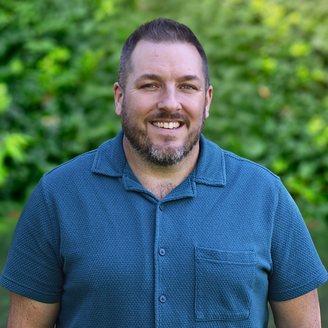 Man with a beard and spiky hair smiles, wearing a blue button-down shirt, against a white wall.