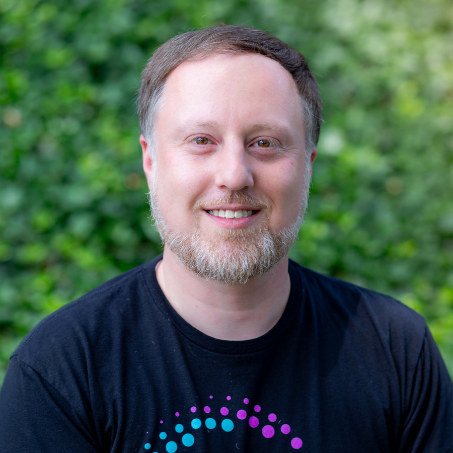 Smiling man with short brown hair and beard in front of green foliage, wearing a black t-shirt with a colorful design.