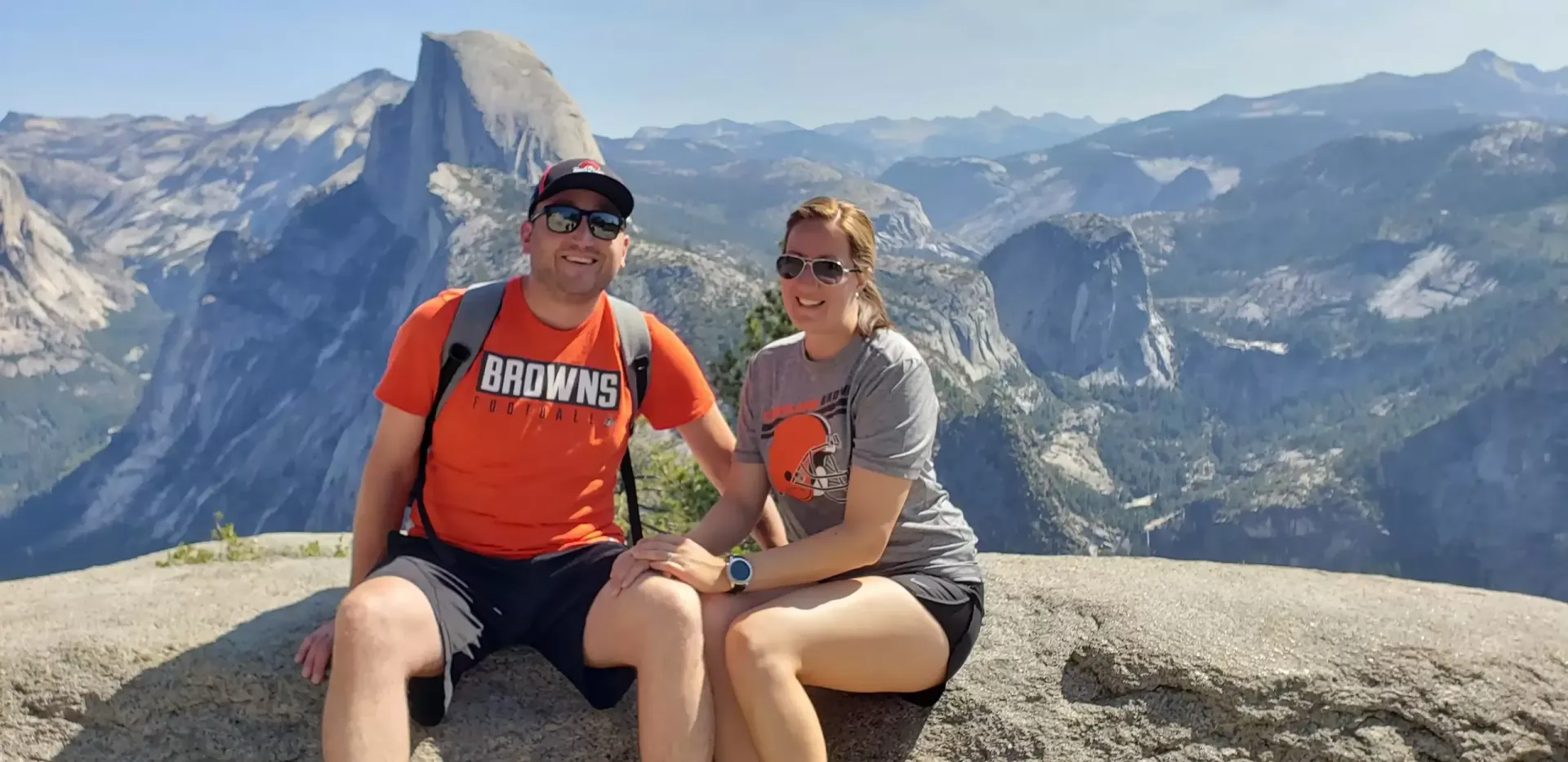 Couple sitting on a rock with mountain landscape in the background. Man wears orange, woman wears gray.
