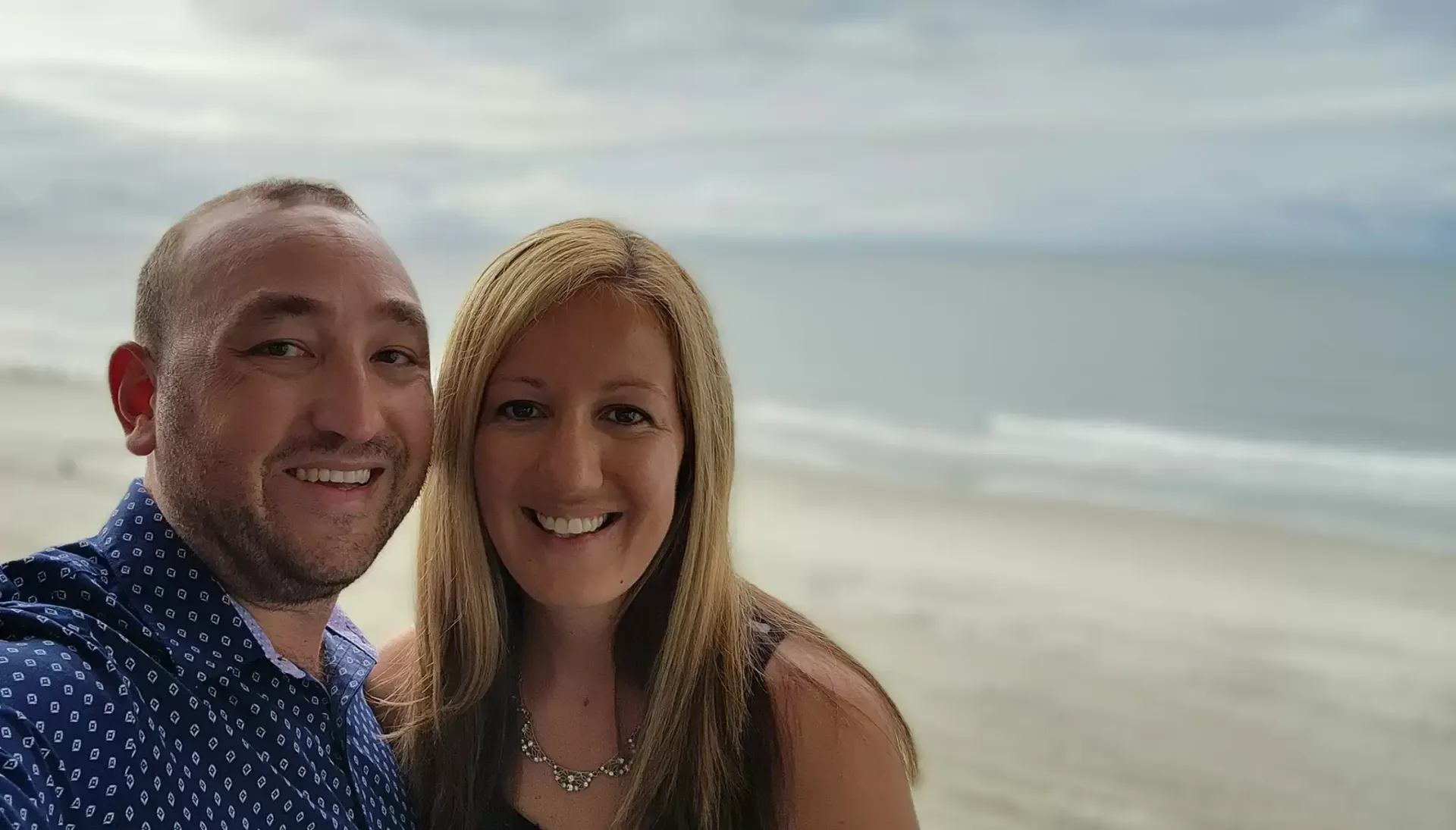 Couple smiling in front of a beach. Man has short brown hair and a blue shirt, woman has long blonde hair.