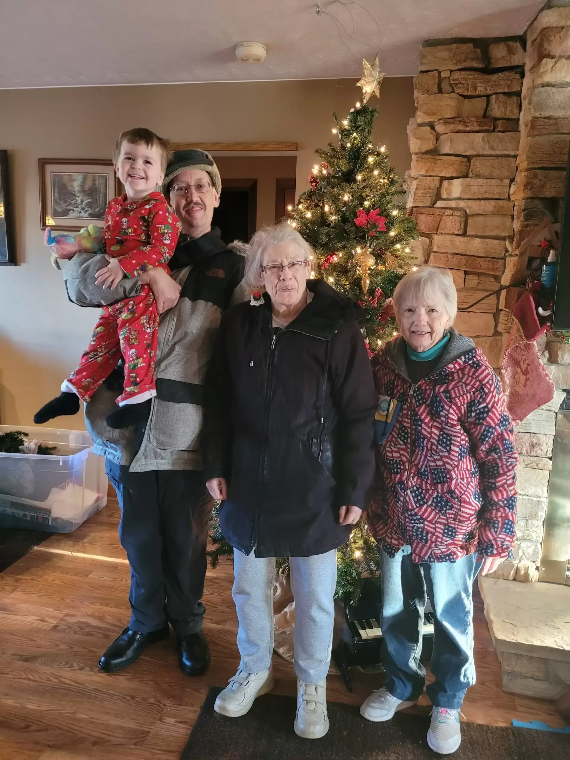 Family poses by Christmas tree. Man holds child in pajamas, two older women smile.
