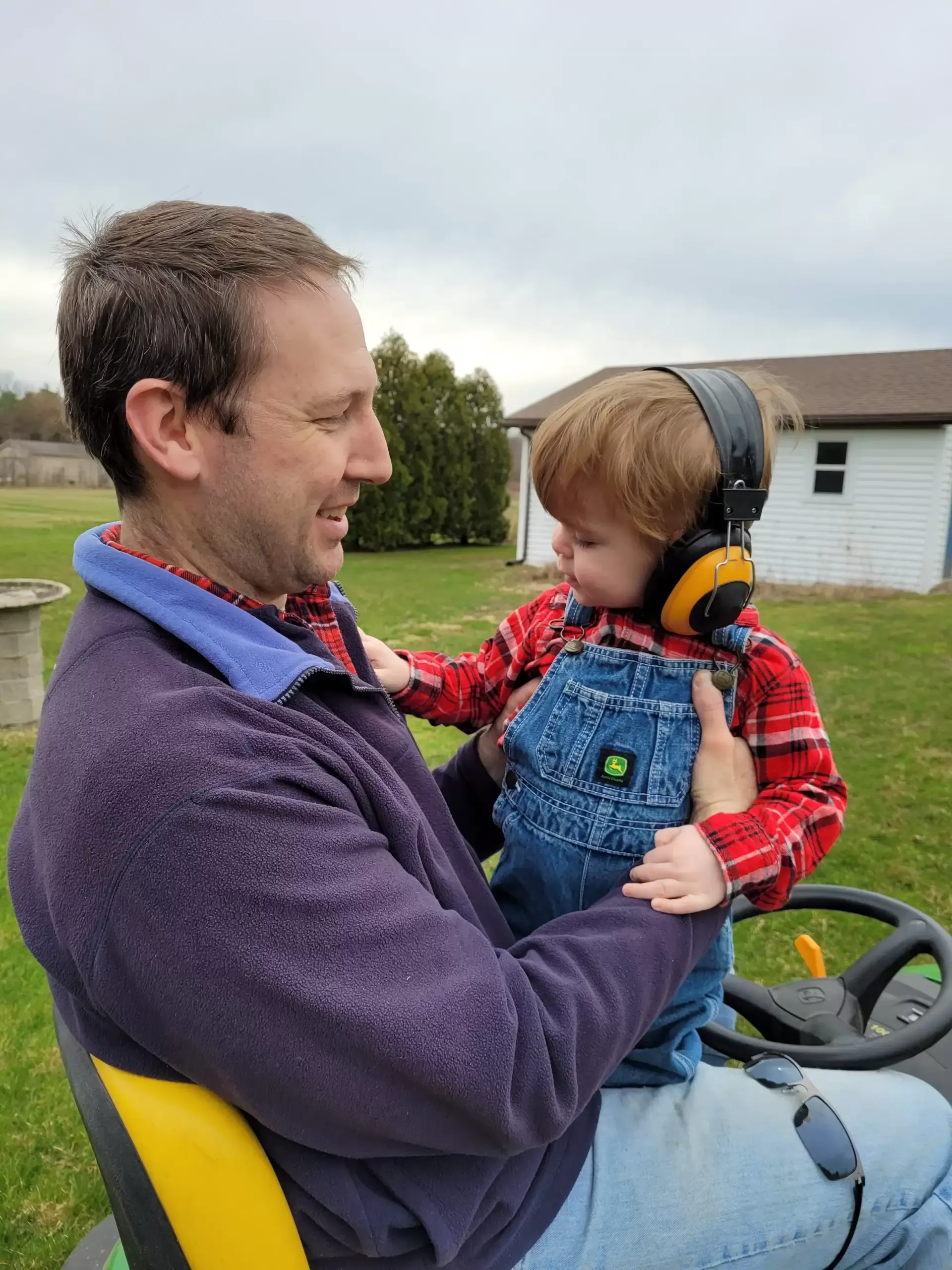 Man holding child wearing earmuffs on a riding lawn mower, outdoors.
