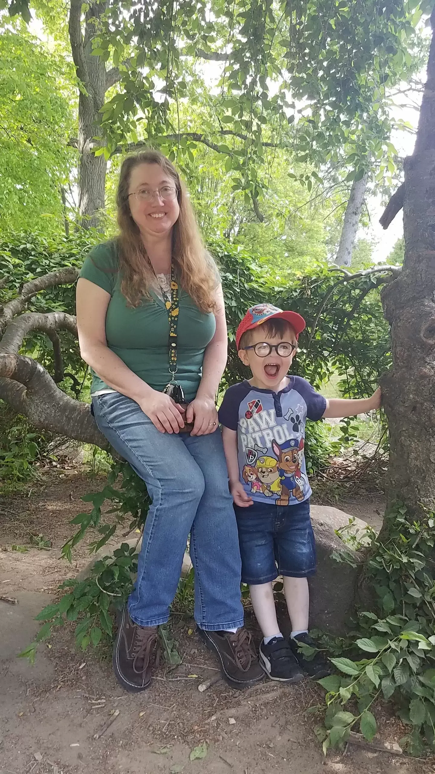Woman and child smiling outdoors, sitting on tree branch. Green shirt, jeans, and hat.