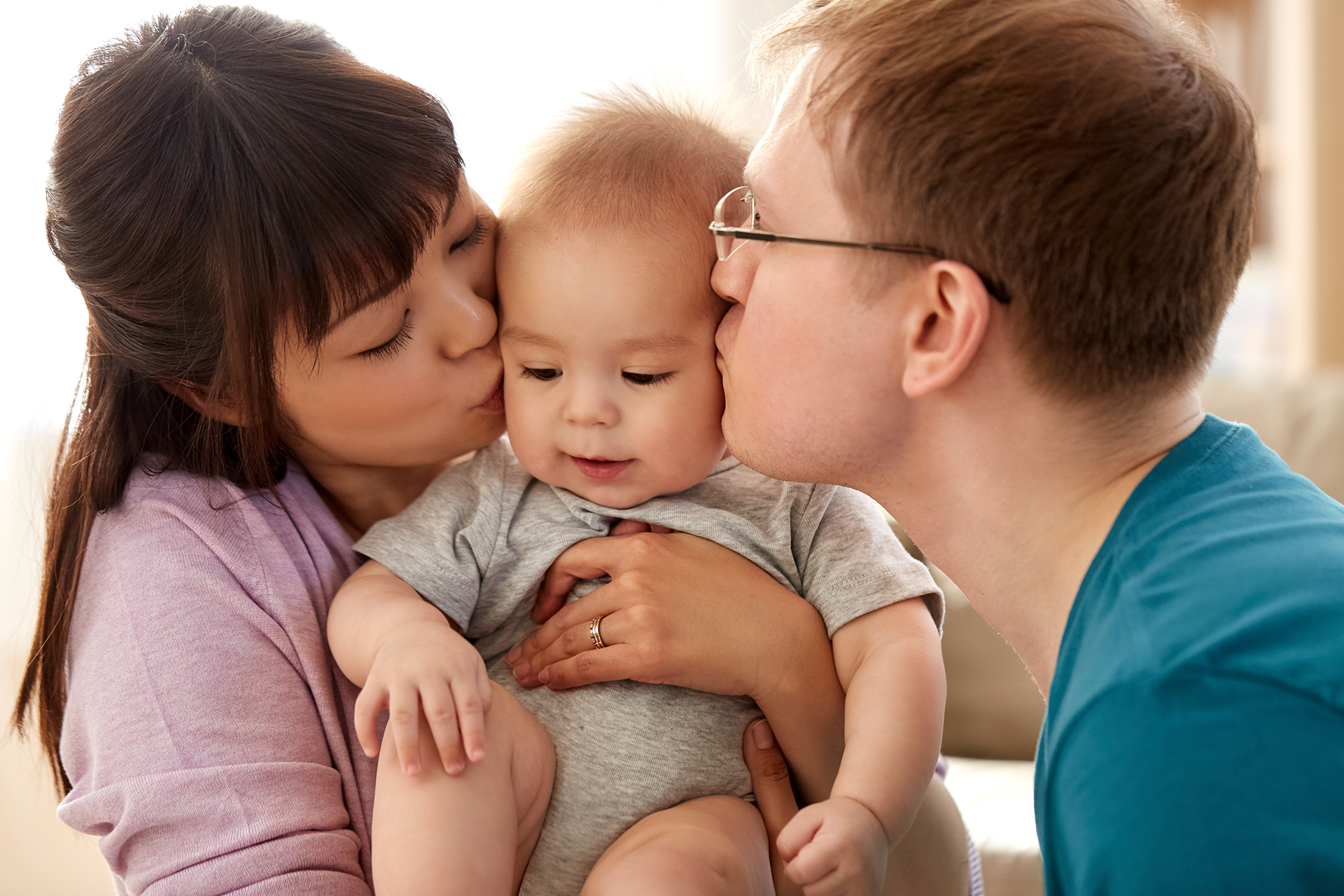 Woman smiles, hugs a baby, both laughing. Baby wears pink headband. Bright, indoor setting.