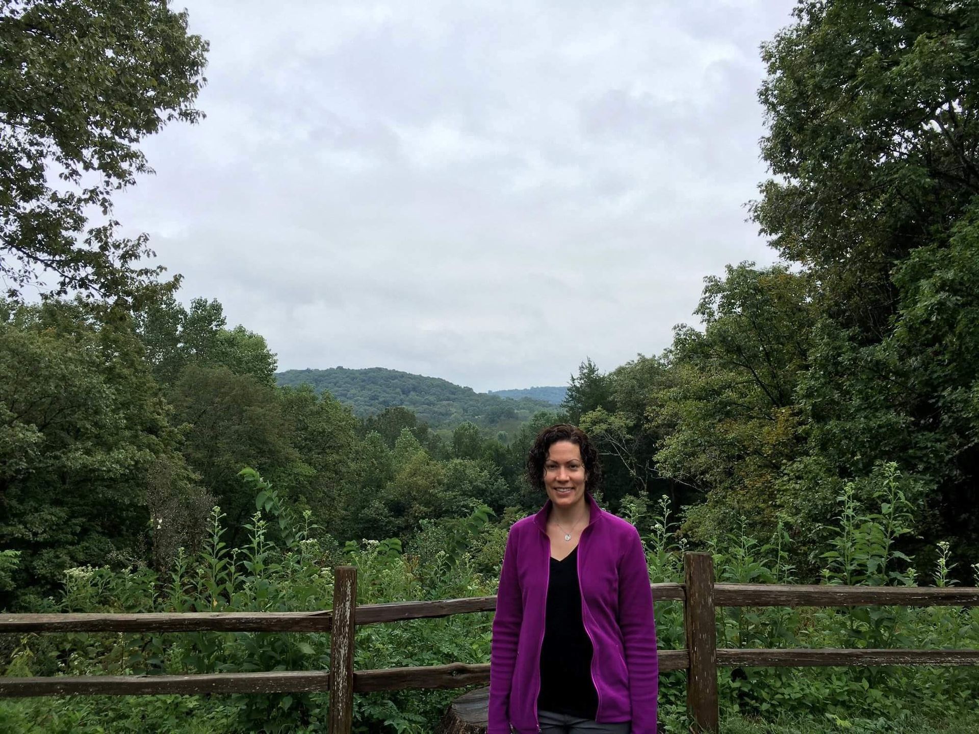 Woman in purple jacket smiles at the camera, standing in front of wooden fence with a green valley and cloudy sky backdrop.