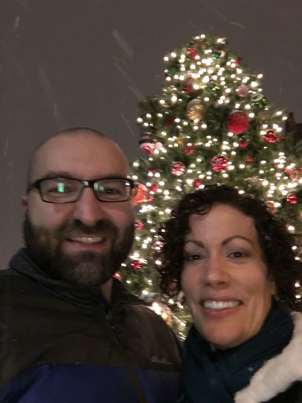 A couple smiles in front of a lit Christmas tree while it snows at night.