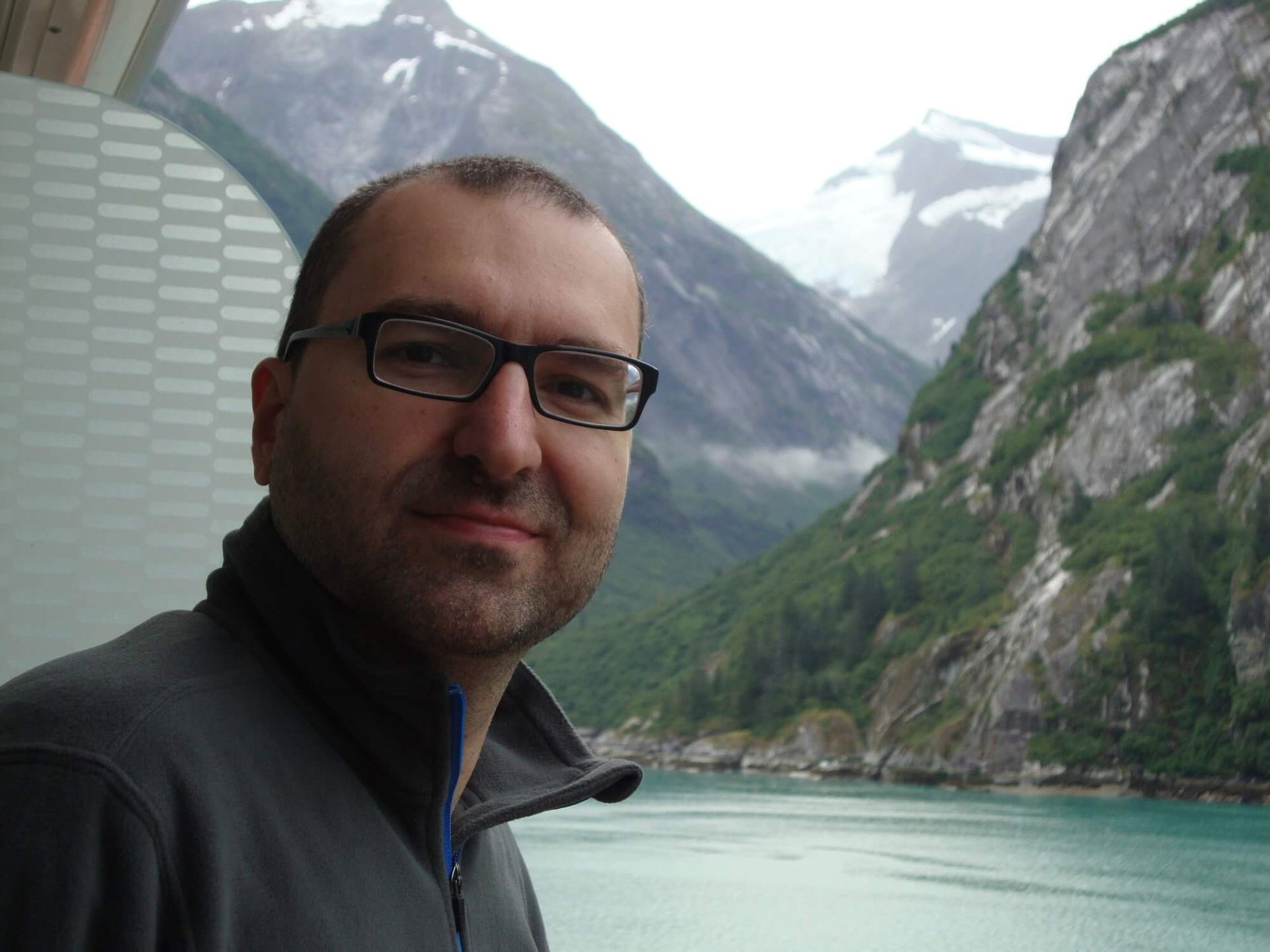 Man with glasses smiling, posing on a balcony overlooking a fjord and mountain range with snow-capped peaks.