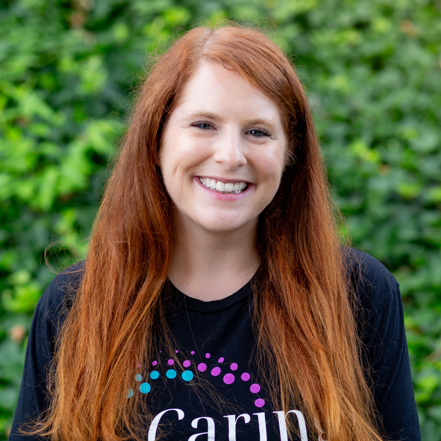 Woman with red hair smiling, wearing a black t-shirt with a logo, in front of green foliage.
