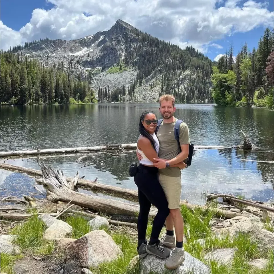 Couple hugs near a lake, with a mountain backdrop. Woman wears white top, black pants; man, t-shirt and shorts.