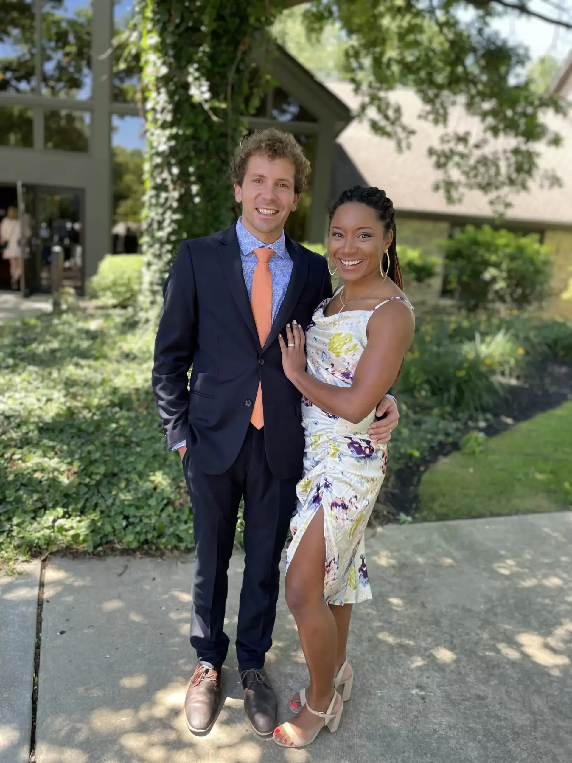 Man in navy suit and woman in floral dress embrace outdoors, smiling.