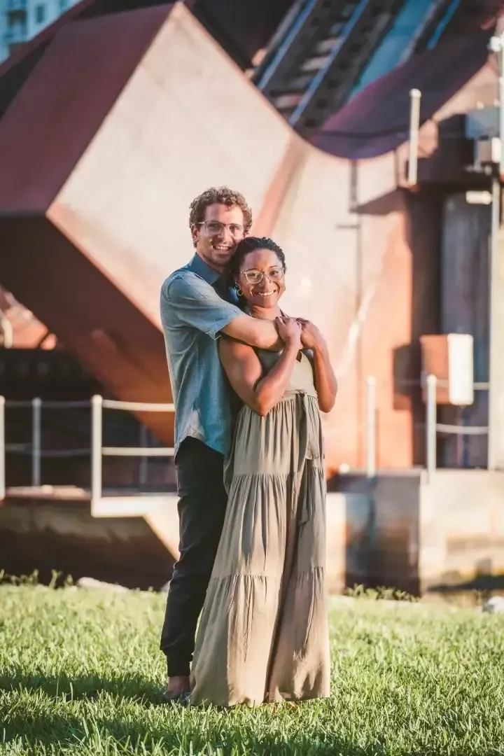 A couple embraces, the man behind the woman. They stand on grass, near a large, rusty metal structure.