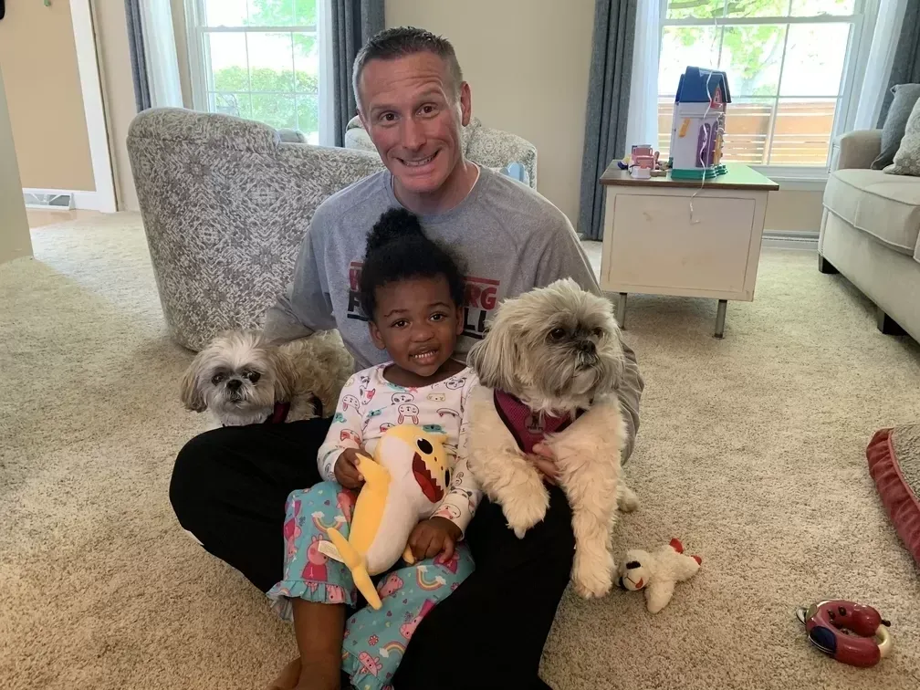 Man and child seated on floor with two dogs. The child holds a toy.