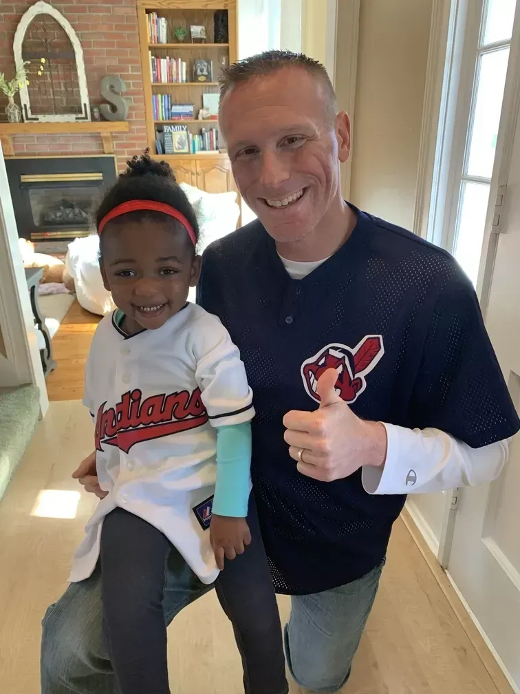Man with a child, both in Cleveland Indians gear, posing indoors. Man gives a thumbs up.