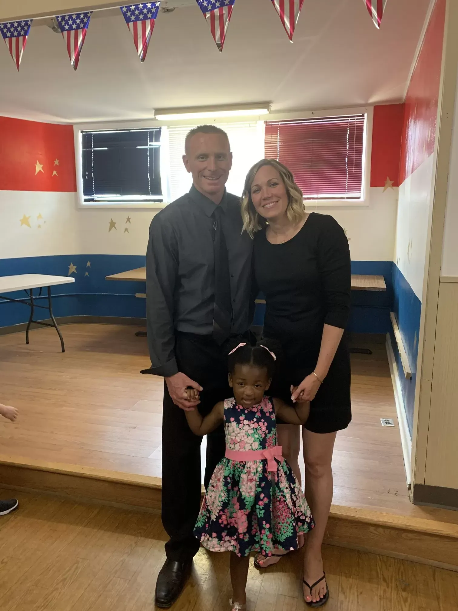 Family of three smiling, holding hands in a room with patriotic bunting and red and blue walls.
