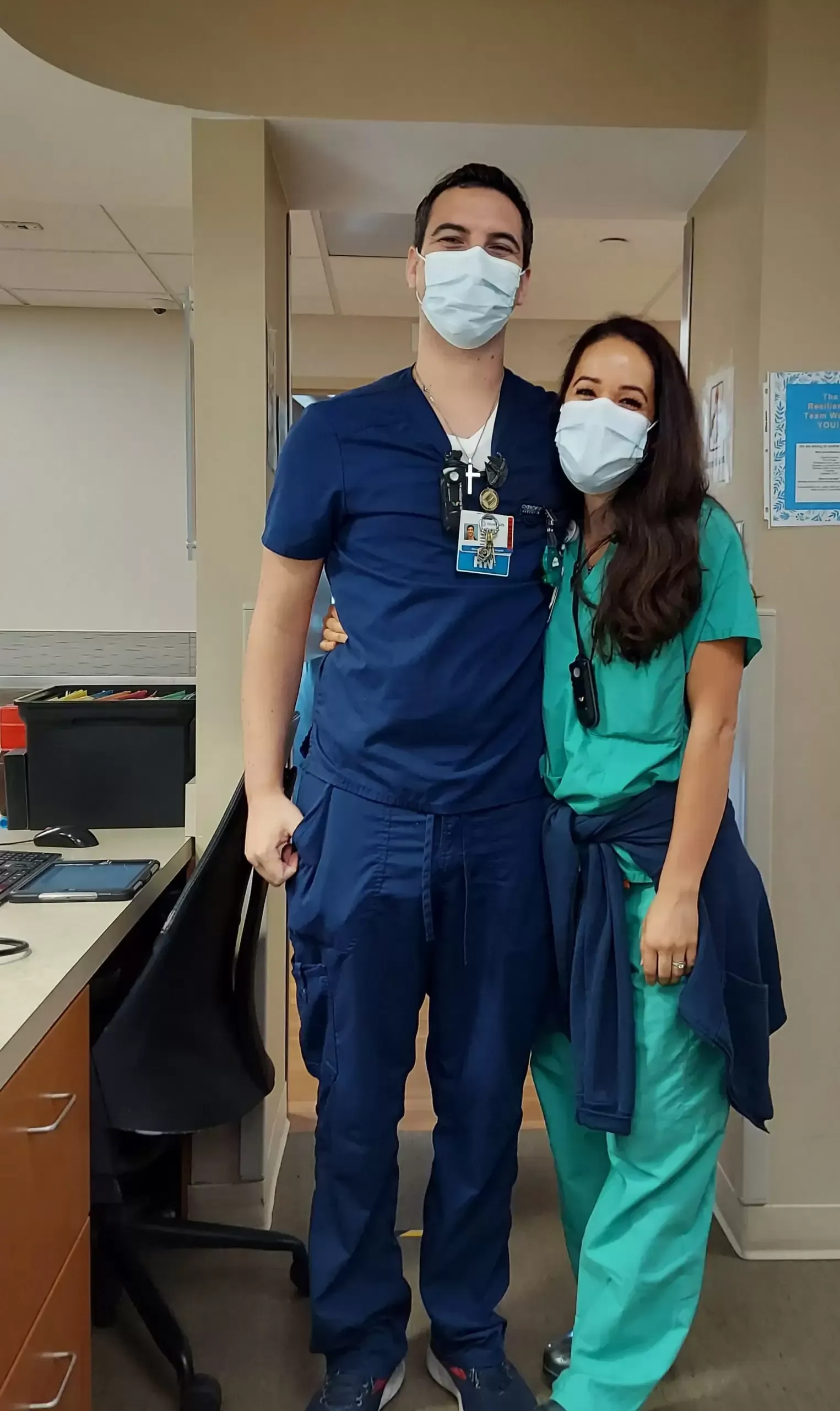 Two masked healthcare workers pose together in a hospital hallway.