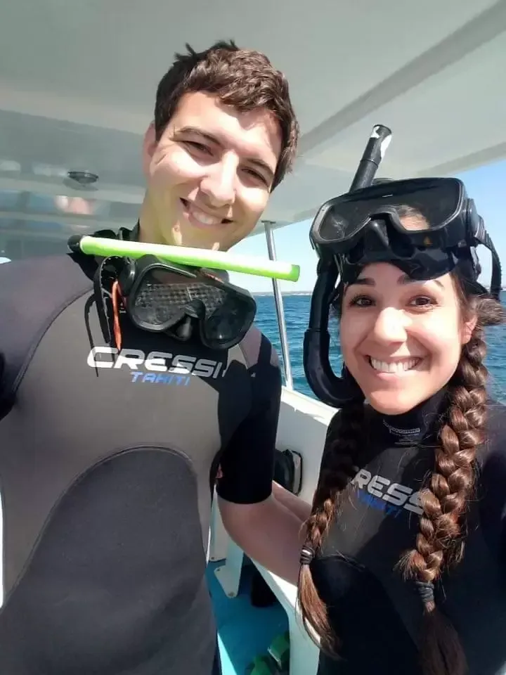 Couple in scuba gear on a boat, smiling at the camera, ocean in the background.