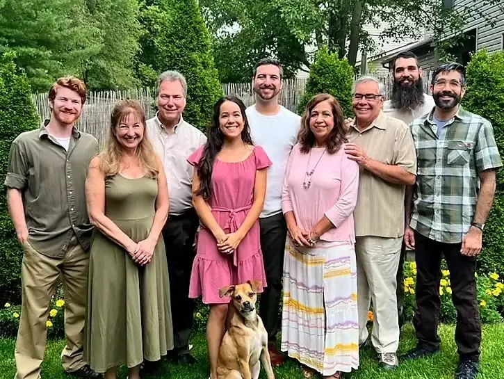 Family group posing outdoors, smiling. Includes a dog. Green and beige tones, lush garden.