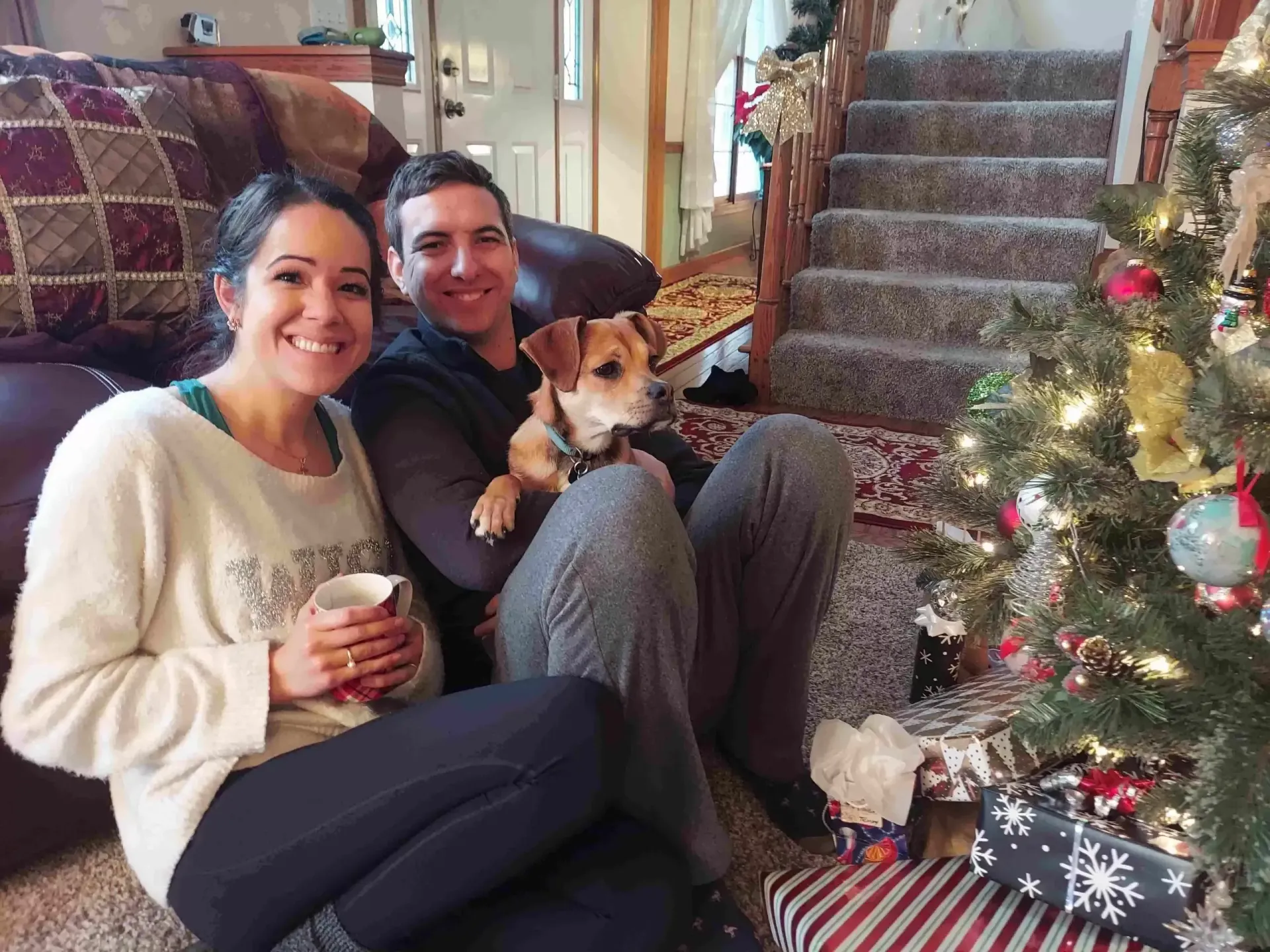 Woman and man sit with a small dog by a Christmas tree; they smile at the camera.